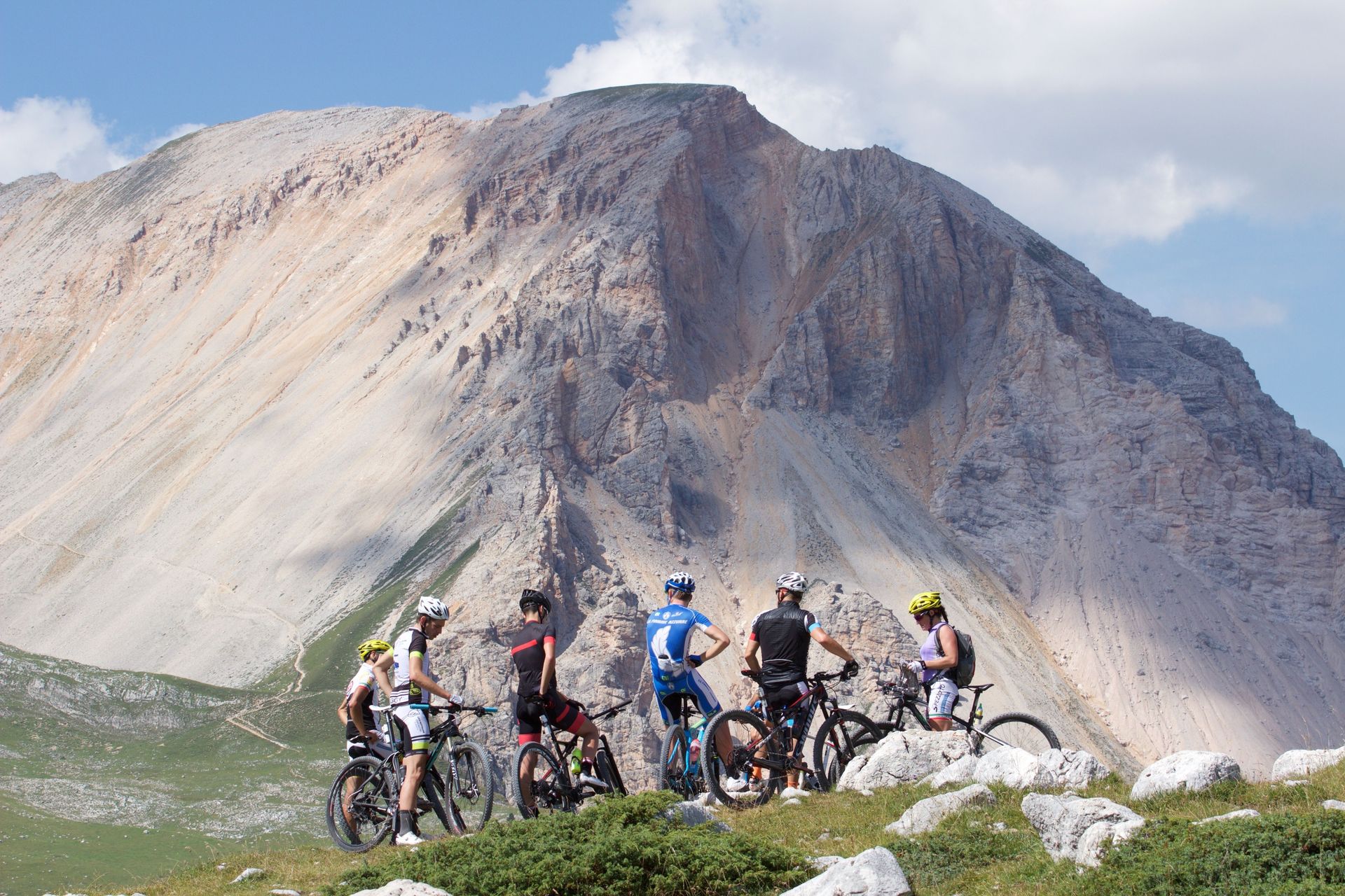 Cyclists gathered with bikes at the base of a rocky mountain slope under a cloudy sky