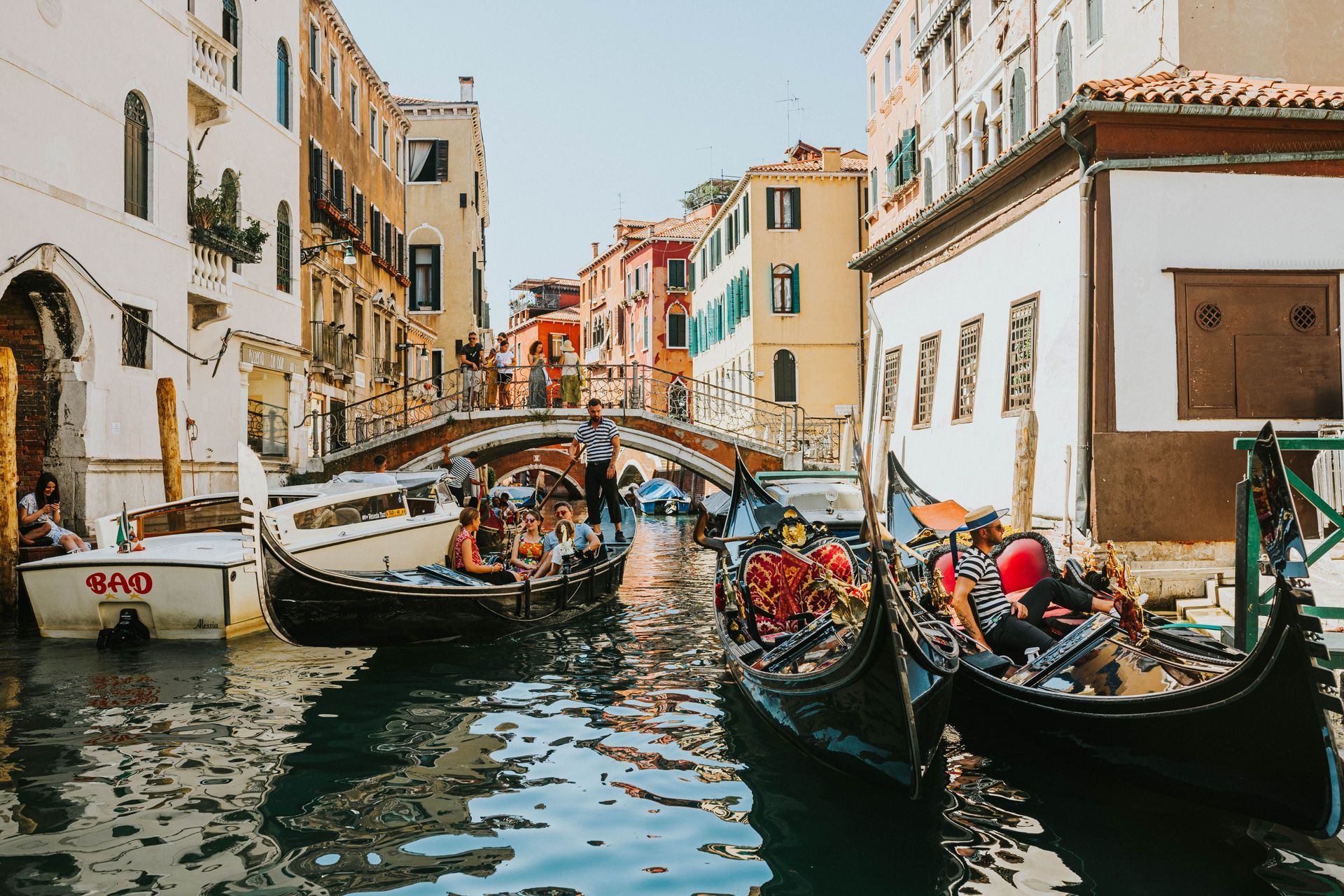 Gondolas on a narrow Venice canal between colorful buildings, with people aboard and reflections on the water