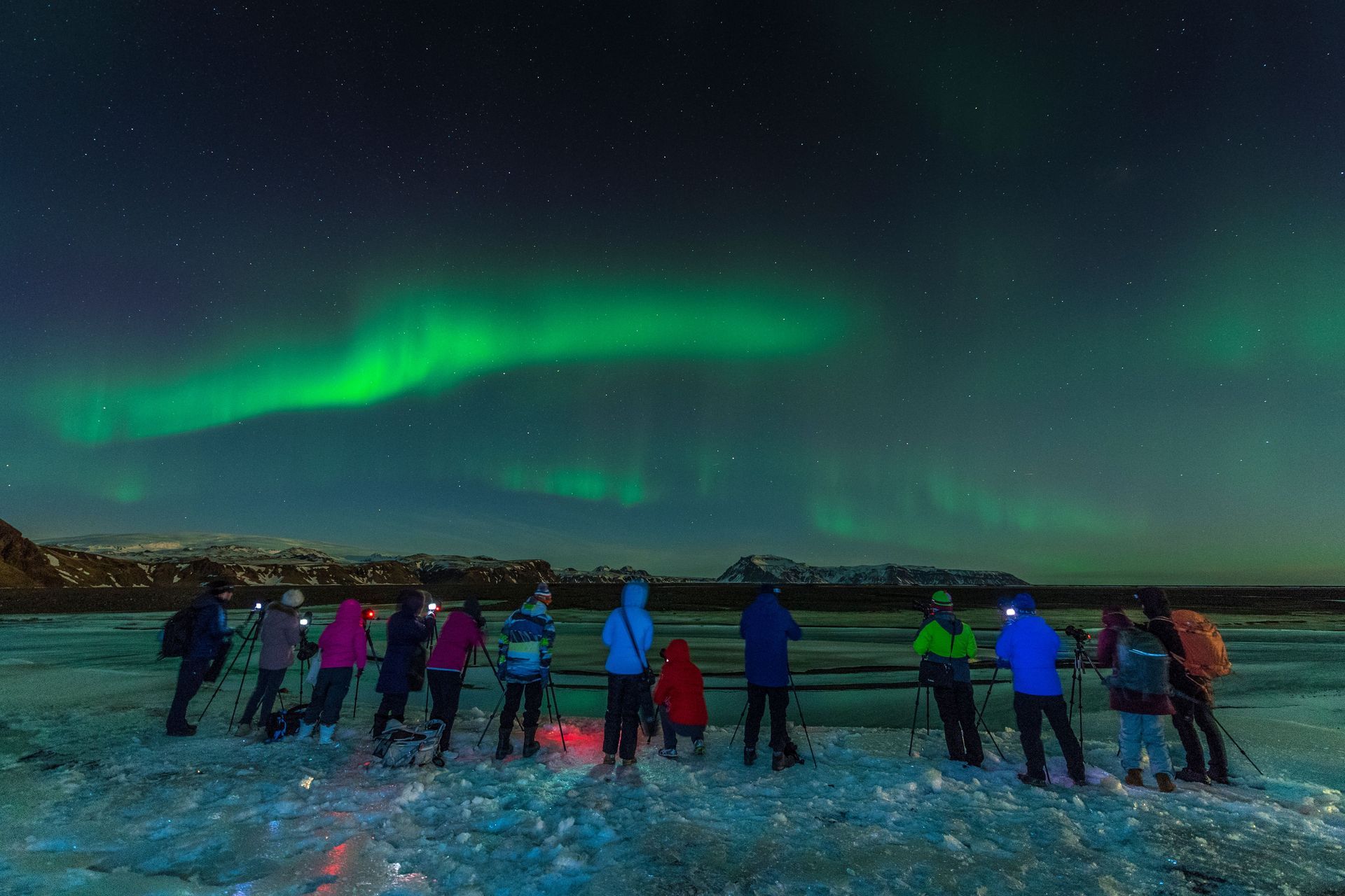 People on a snowy shore watching green northern lights in a dark night sky
