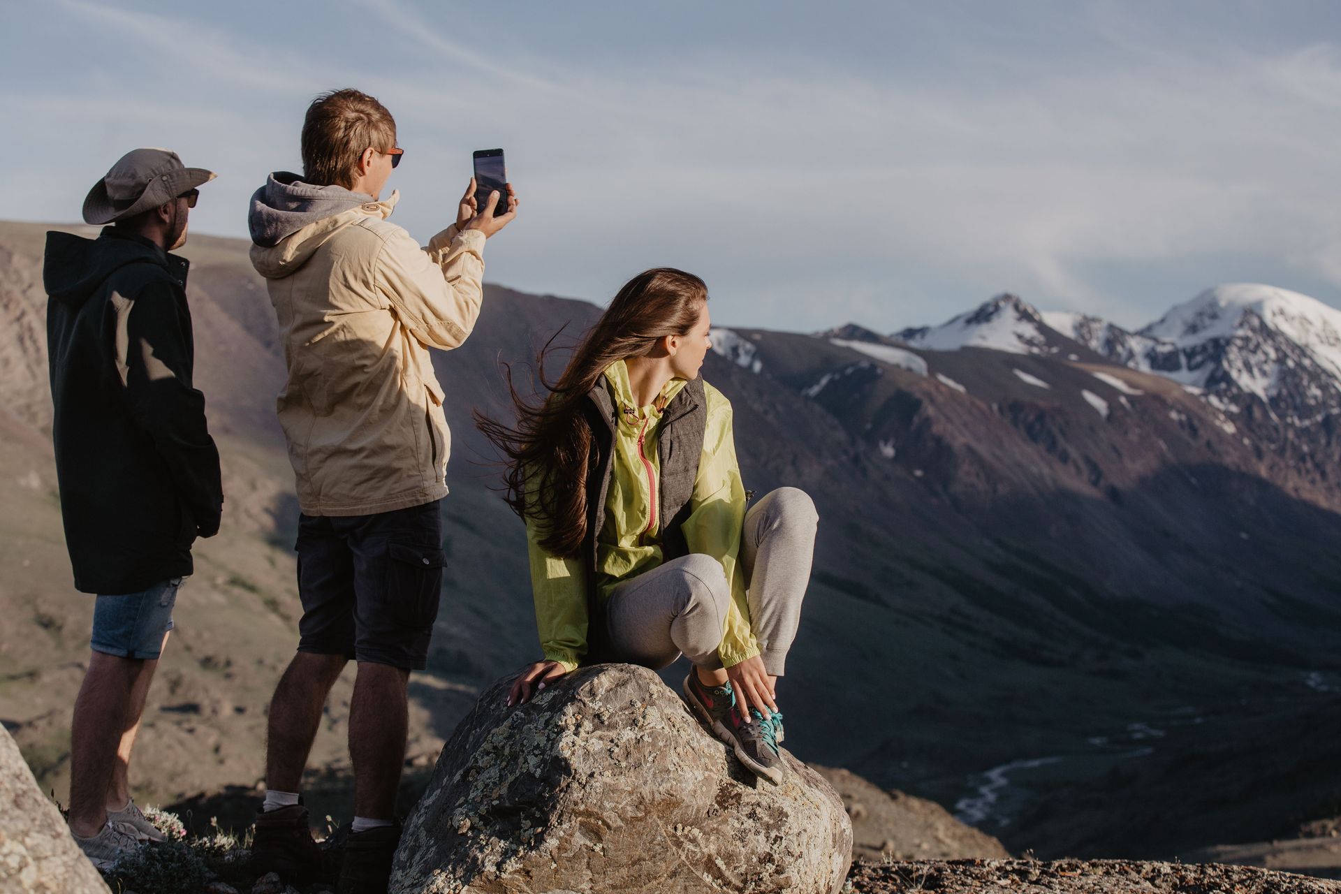 Three hikers on a mountain overlook, one taking a photo with snowy peaks in the background