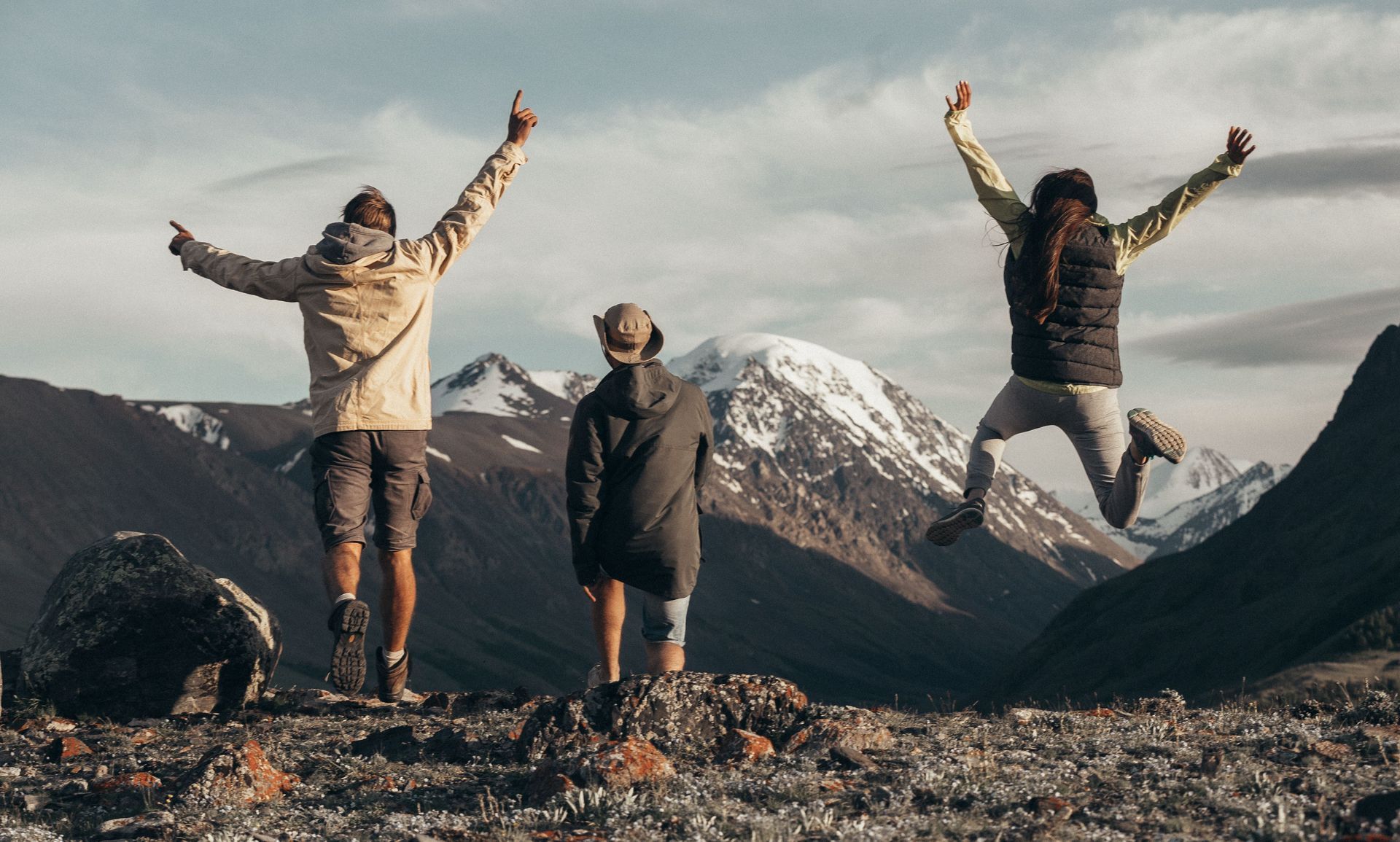 Three hikers jump with arms raised on rocky ground before snowcapped mountains at sunset