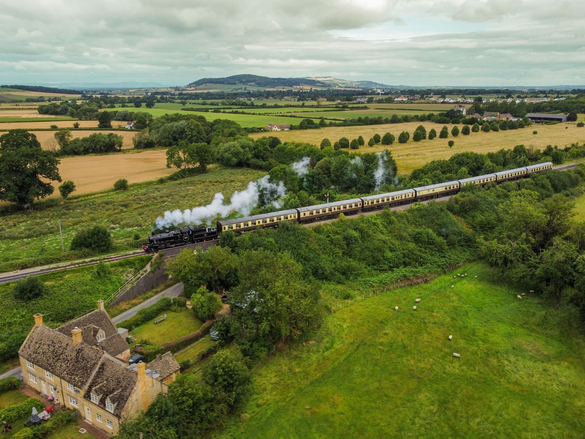 Steam train crossing a rural viaduct amid green fields and trees, with smoke trailing behind.