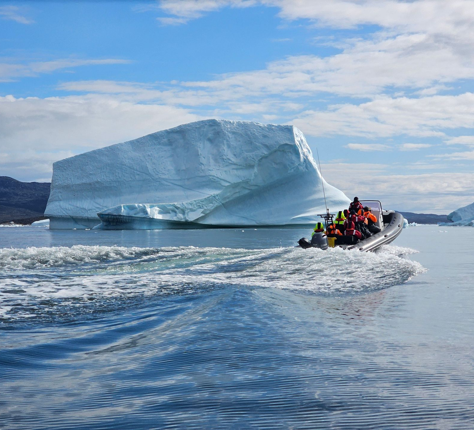 Tour boat with people near a massive blue iceberg on calm water under a cloudy sky