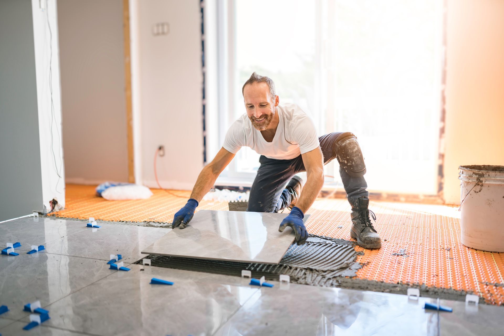Man installing tile flooring in a room, kneeling and smiling, surrounded by tools and materials.