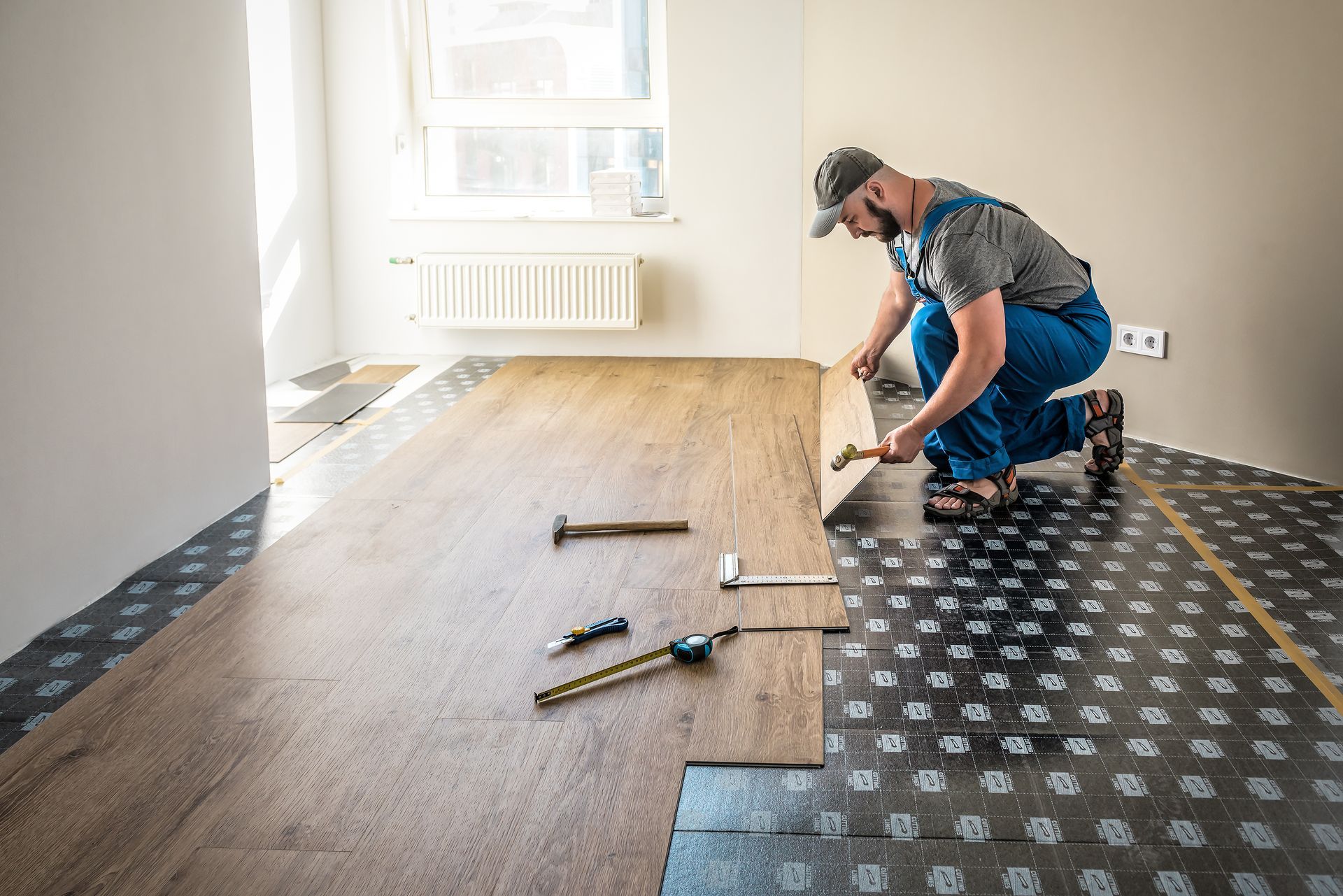 A man installs wood flooring, using a hammer and other tools, in a room with a window and light-colored walls.