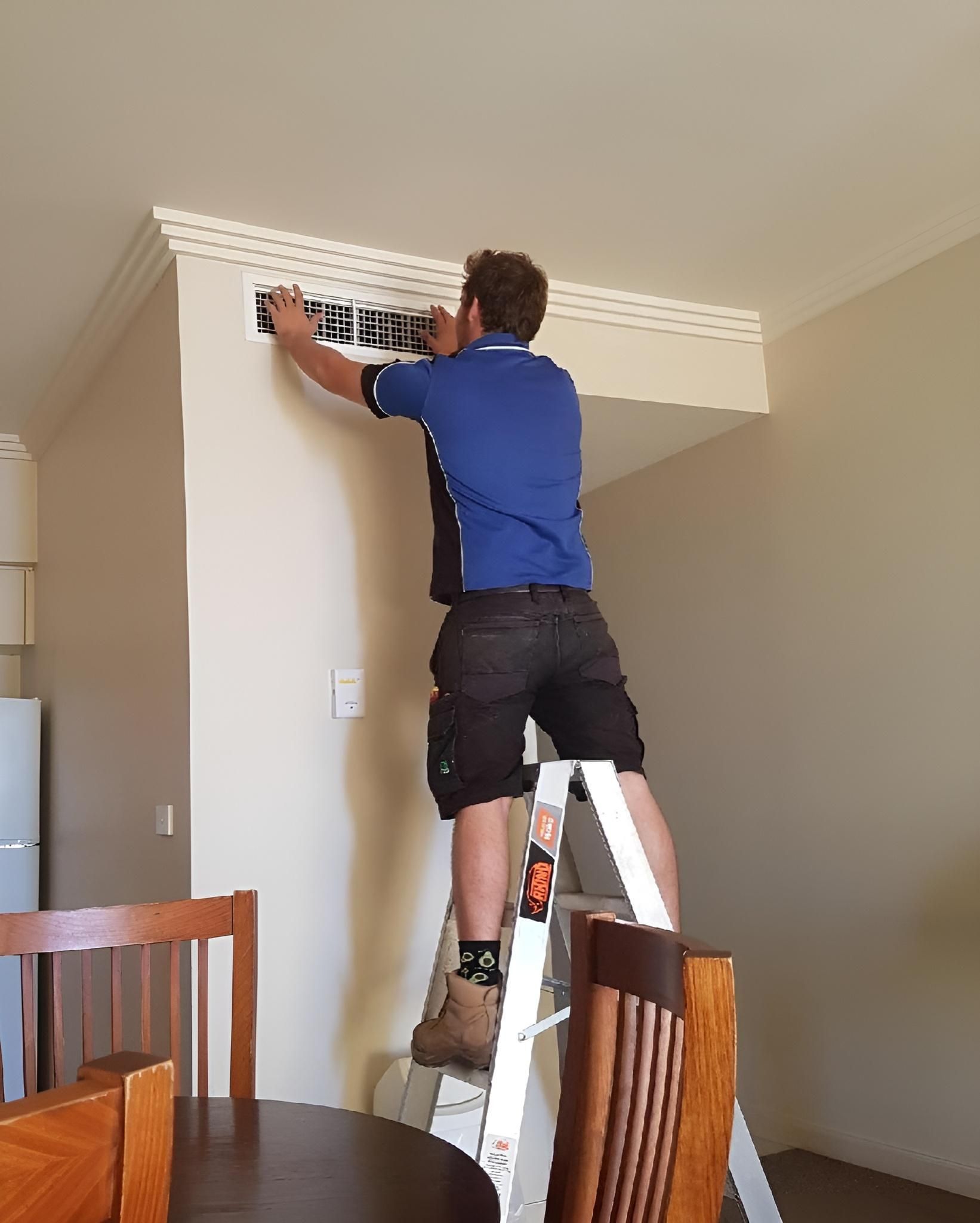 A Man is Standing on a Ladder Fixing a Vent on a Wall — Atmosphere Air Conditioning & Refrigeration In Taylors Beach, NSW