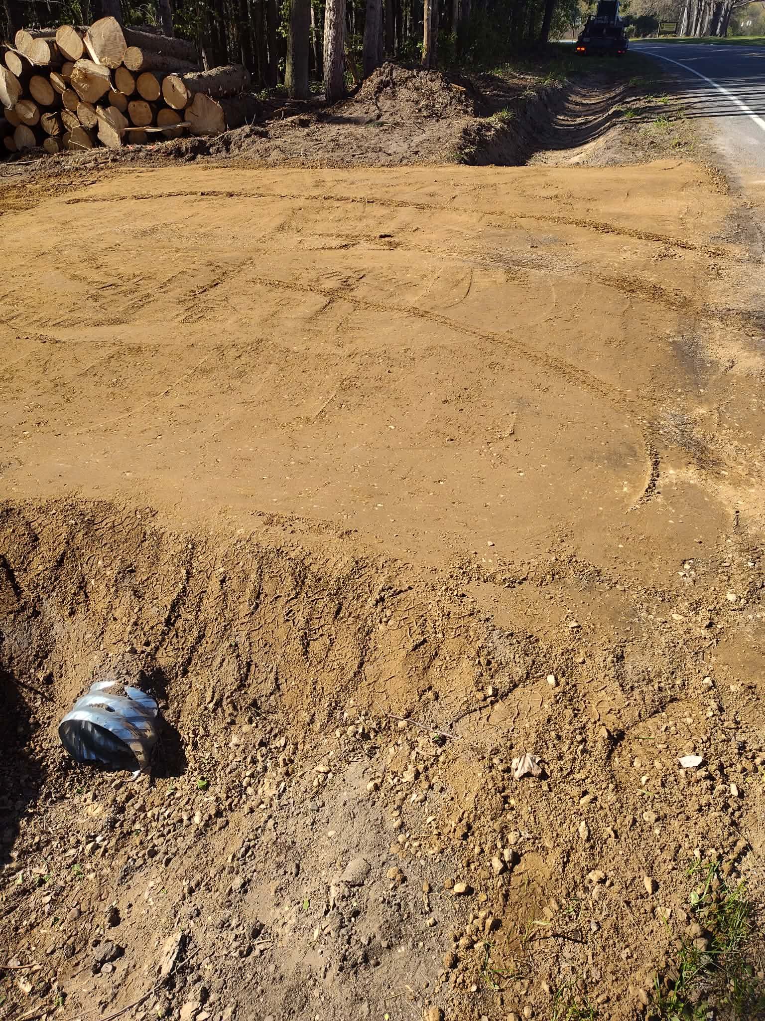 Freshly graded dirt driveway beside stacked logs and trees, with a small metal cup in the foreground