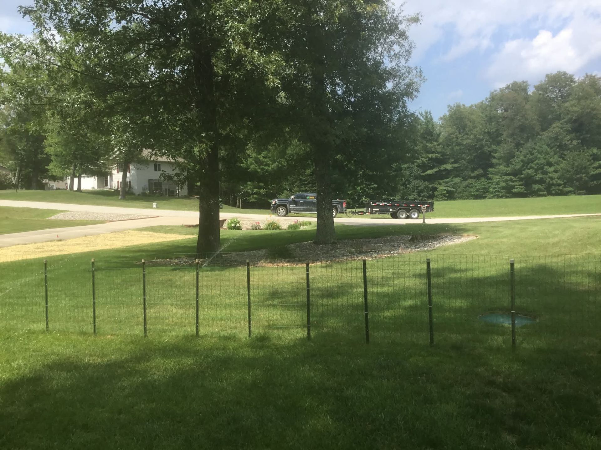 Grassy park with trees, a fence, and a road with parked cars under a partly cloudy sky
