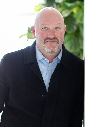 Portrait of a bald man in a dark coat and light blue shirt, standing outdoors with greenery behind him