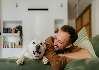 Man lying on a couch hugging a brown dog, both looking happy in a bright living room