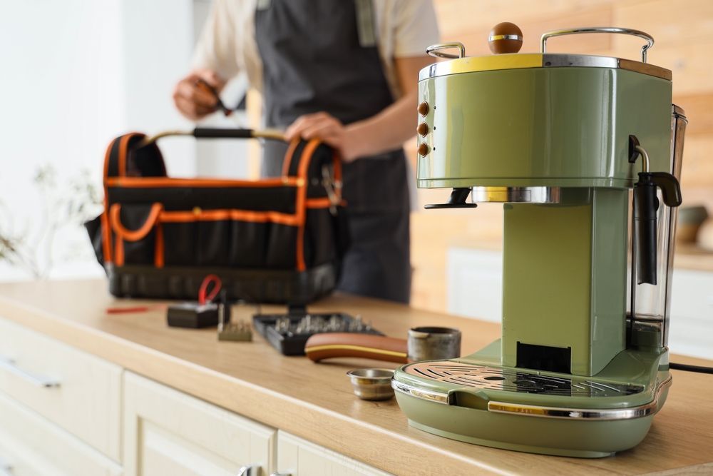 Person repairing a retro green coffee maker on a countertop, with tools and toolbox in the background.
