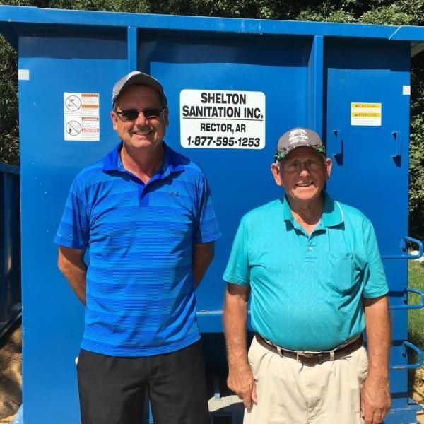 Two men standing in front of a blue Shelton Sanitation Inc. bin dumpster