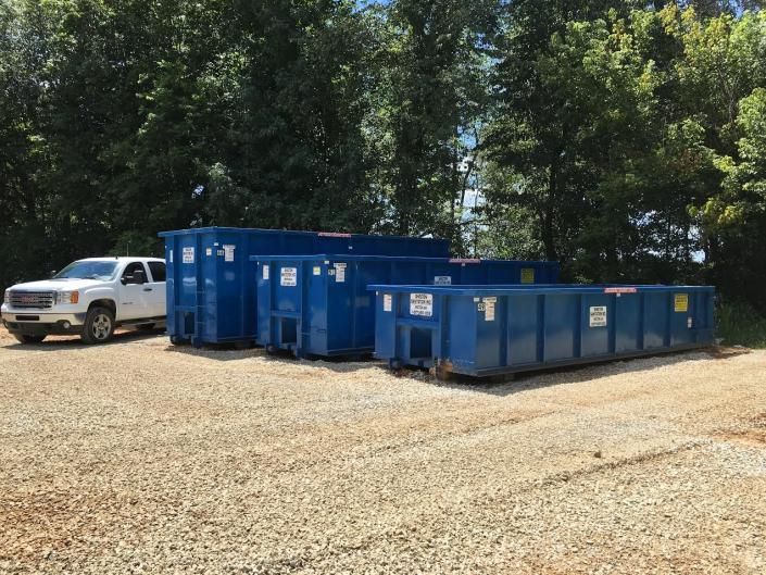 Blue dumpsters beside a white truck on a gravel lot with trees in the background