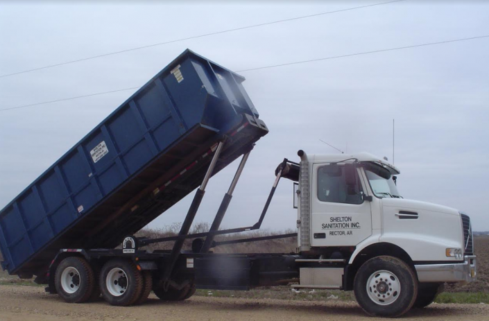 Blue dump trailer raised on a white truck, unloading at a roadside site.