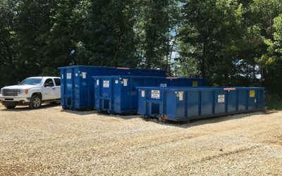 Blue dumpsters lined up on a gravel lot beside a white pickup truck and trees