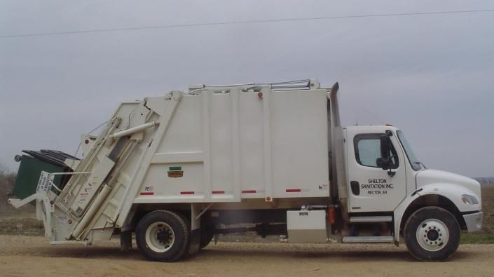 White garbage truck viewed from the side on a dirt road under a cloudy sky.