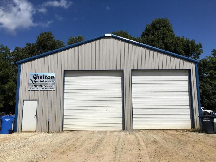Gray metal garage building with two white roll-up doors and a small office door on the left.