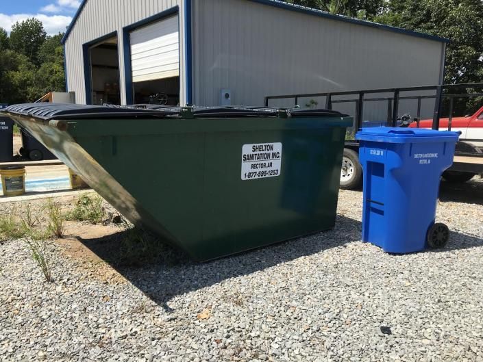 Green dumpster and blue recycling bin on gravel outside a warehouse