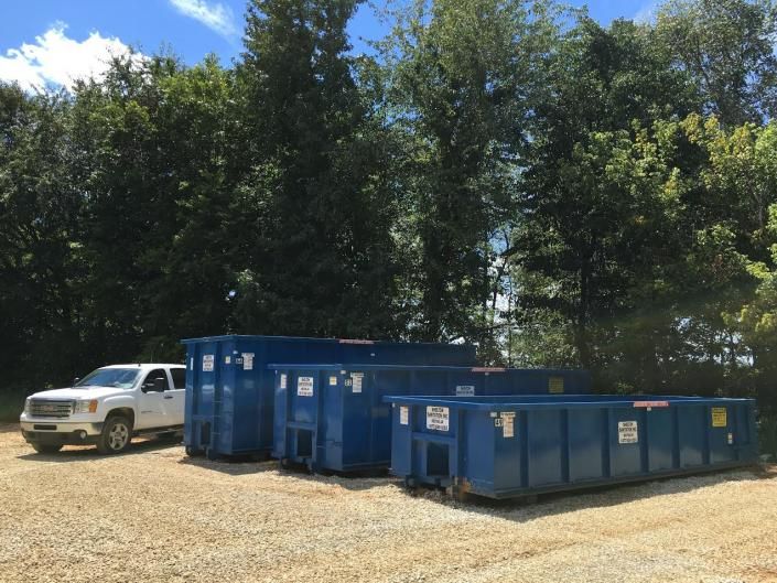 White truck beside blue dumpsters in a wooded gravel lot under a bright sky