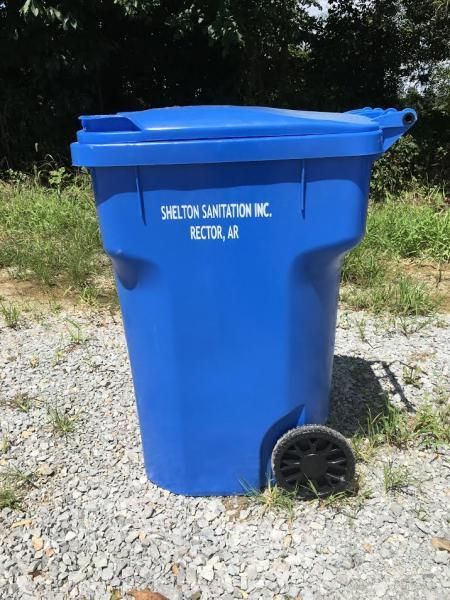 Blue rolling trash bin labeled “Shelton Sanitation, Inc.” on a grassy roadside.