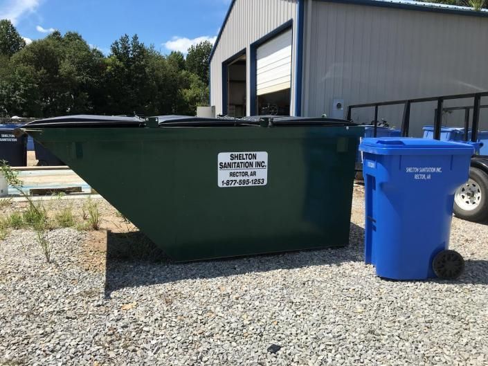 Green dumpster and blue recycling bin outside an industrial building on a gravel lot.