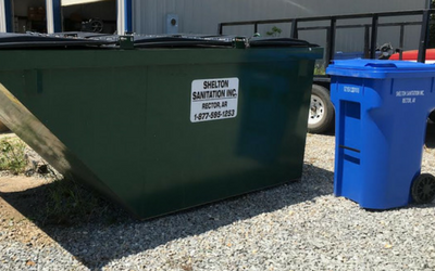 Large green dumpster beside a blue recycling bin on a gravel lot outdoors