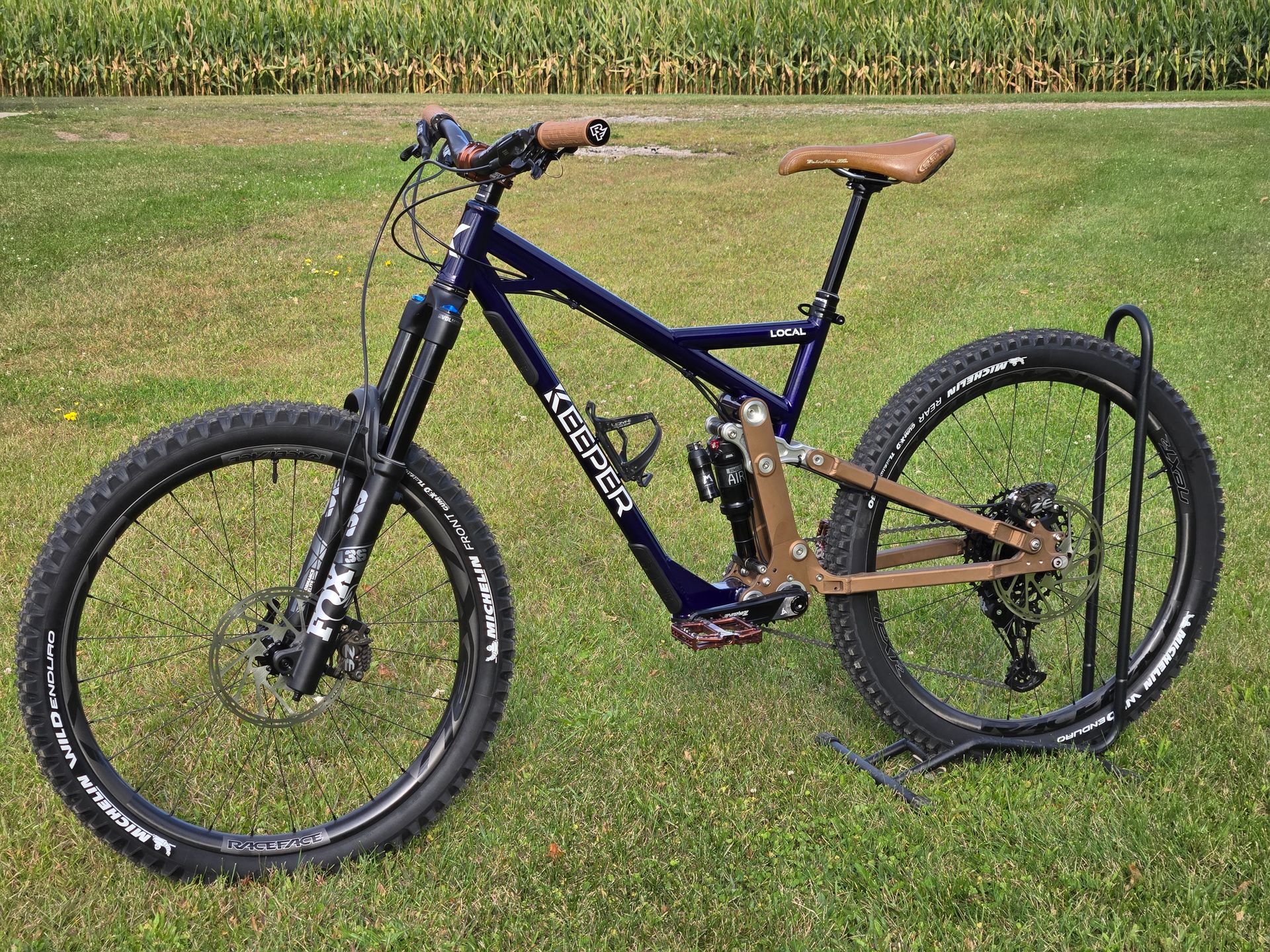 Black mountain bike on a forest trail, leaning against a tree, with brown handlebars and tan grips.