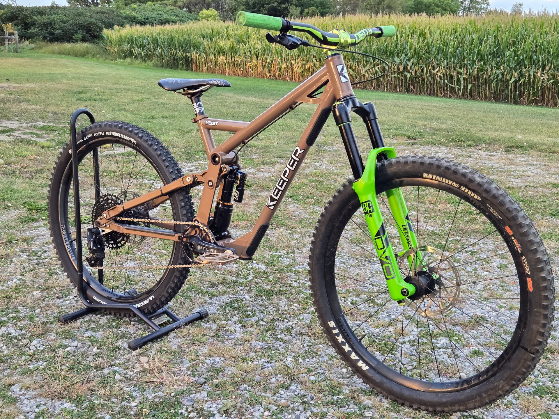 Brown mountain bike with green accents on a stand, in front of a cornfield.