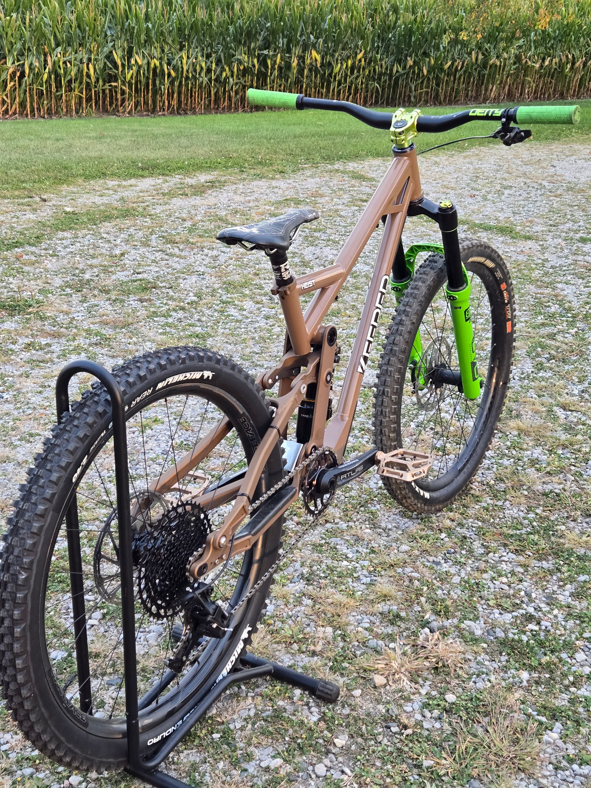 Brown mountain bike on a stand with green accents, outdoors on gravel.