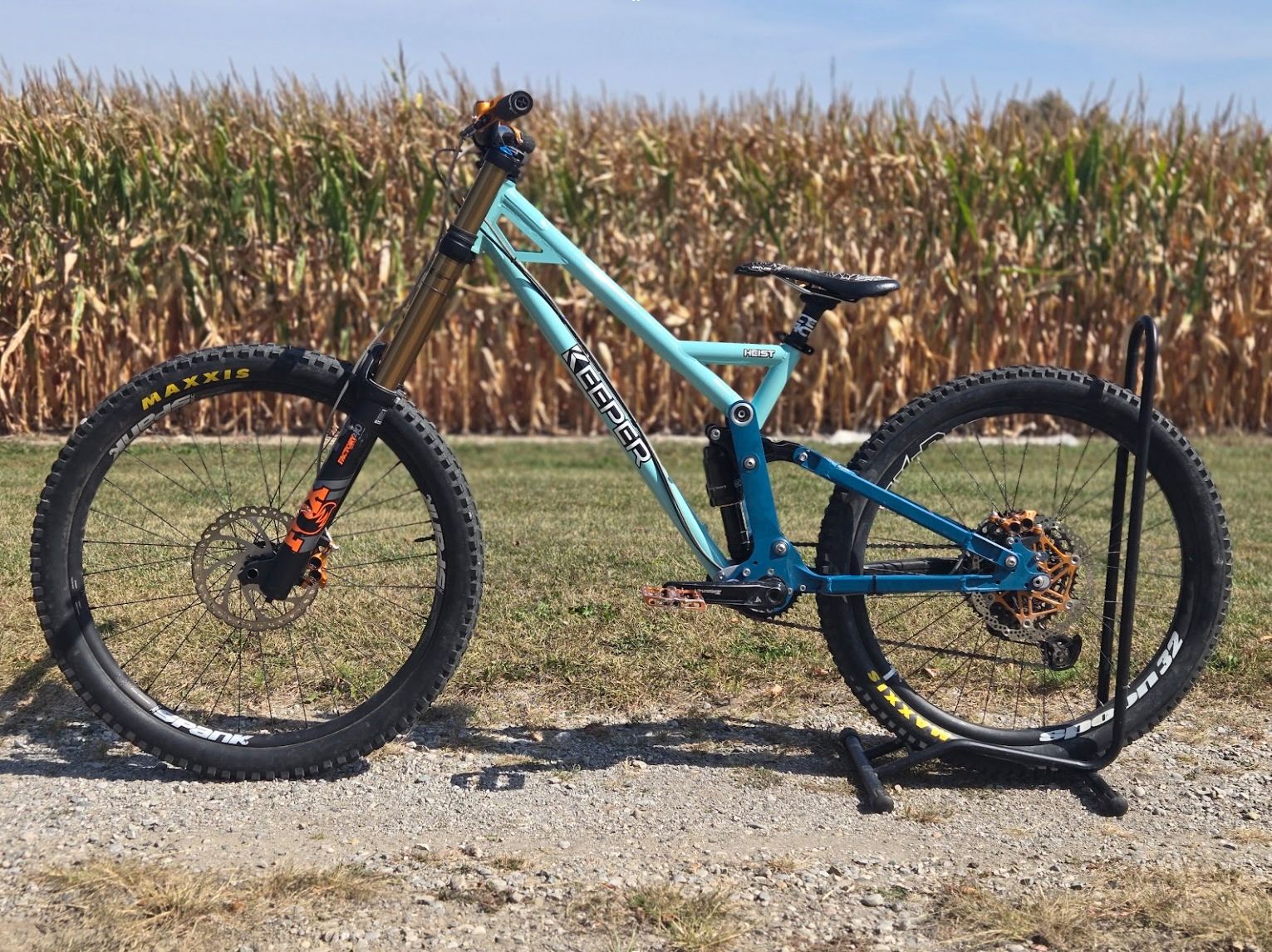 Light blue and teal mountain bike on a stand, front fork and rear hub are gold, field background.