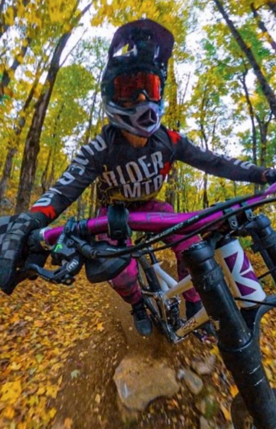 Mountain biker wearing a helmet and protective gear, riding a bike on a trail in a forest with autumn leaves.
