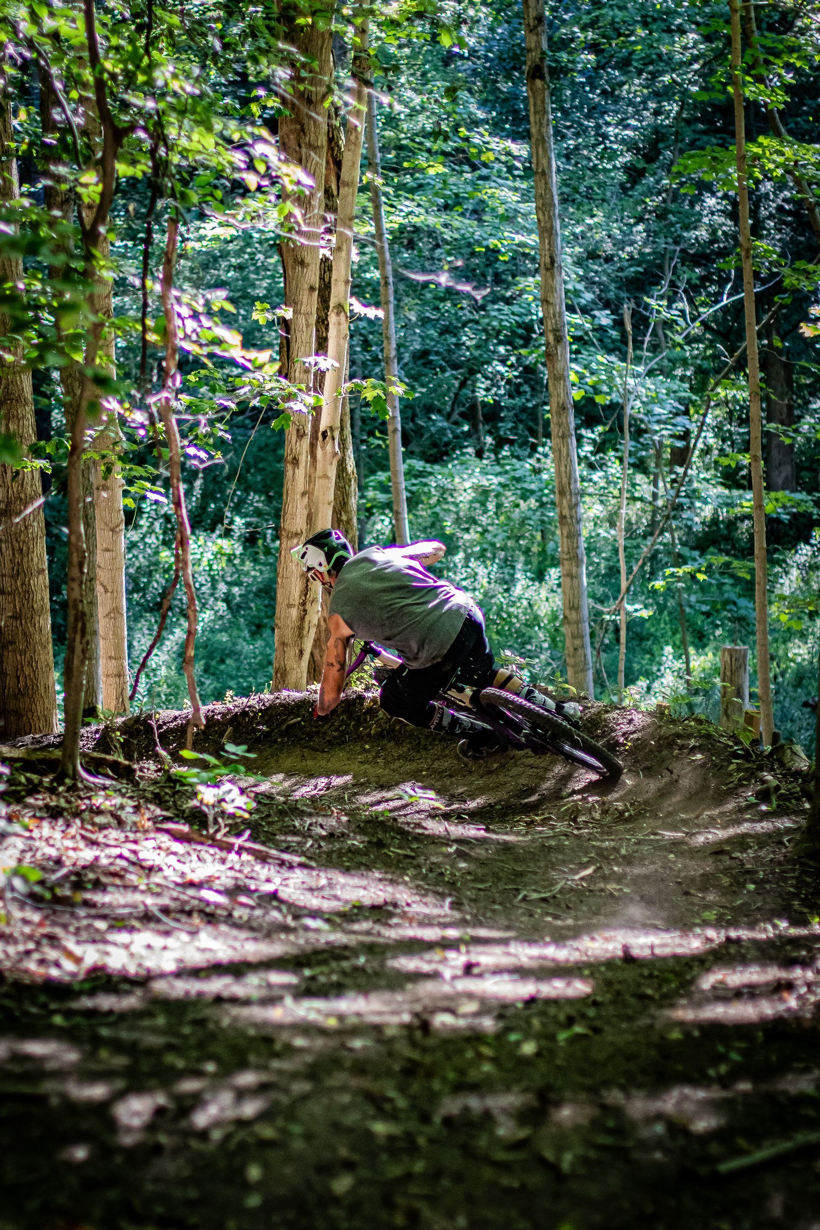 Mountain biker in mid-air over a jump on a wooded trail. Another biker watches. Trees surround them.