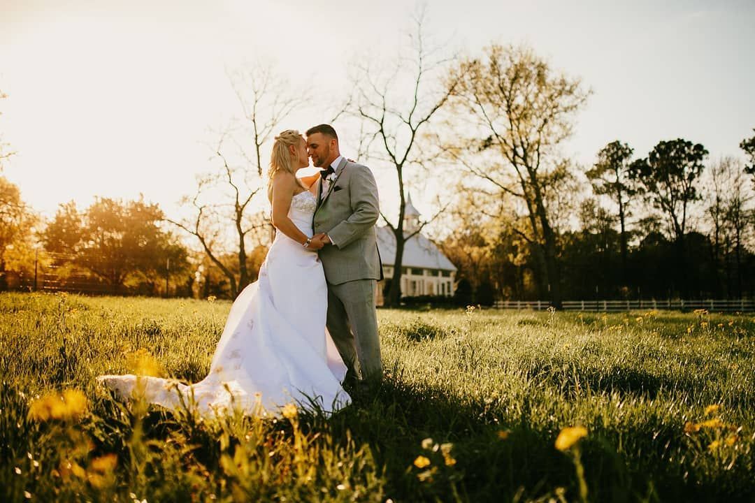 Bride and groom in wedding attire embrace in a field, sun setting behind them.