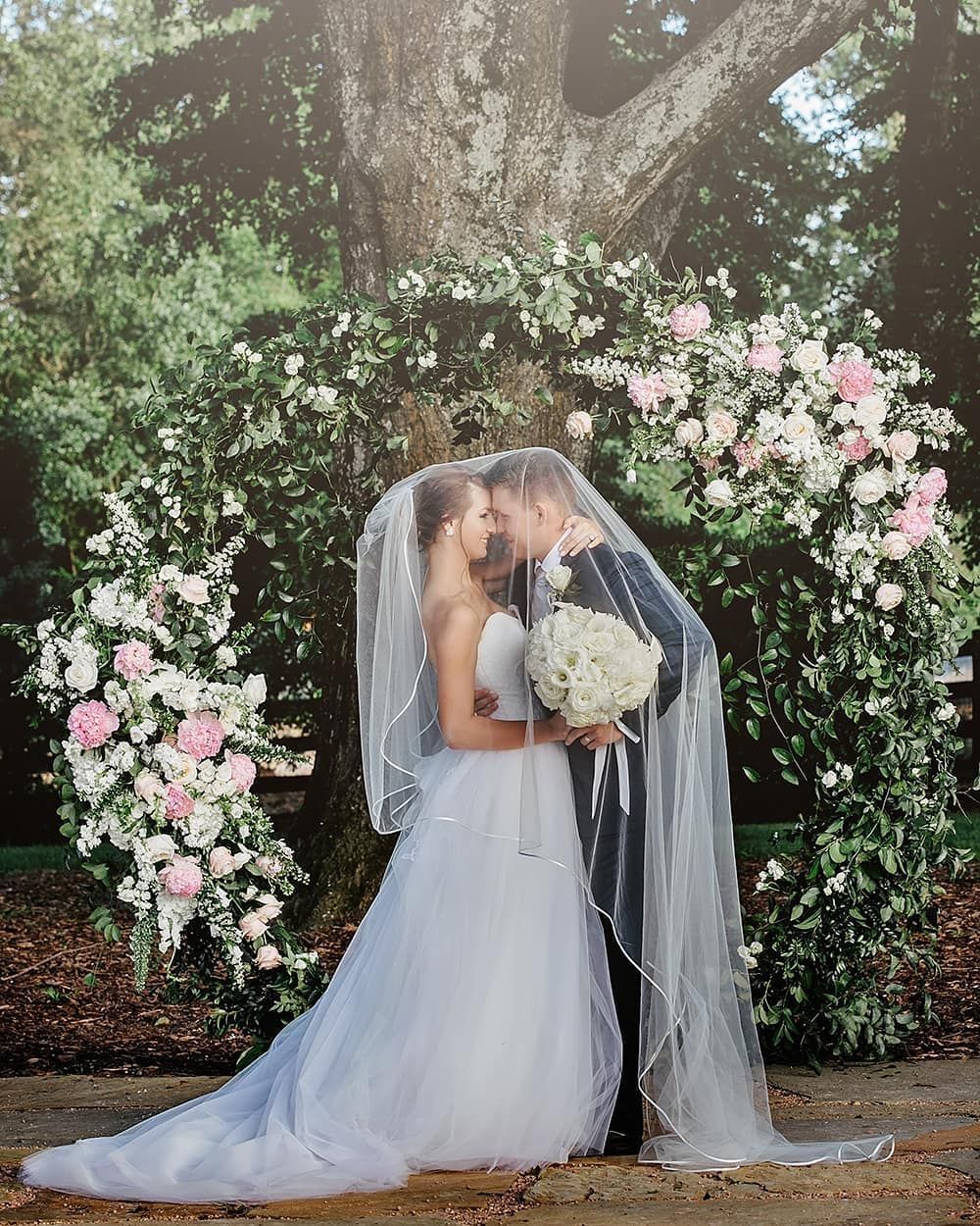 Bride and groom kissing under veil, floral archway. Formal outdoor wedding ceremony.