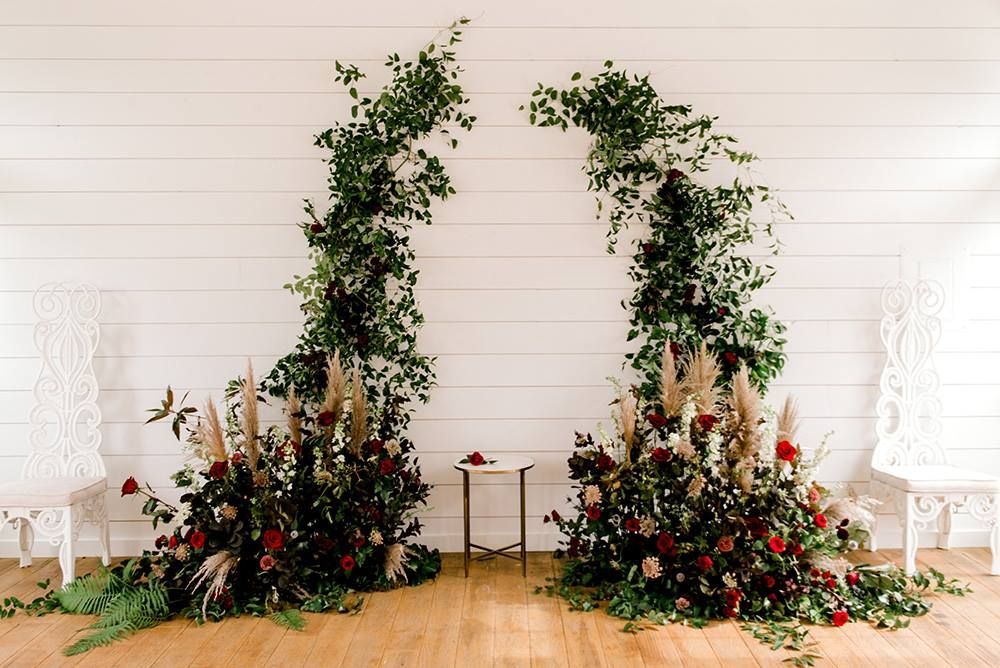 Floral wedding arch with red roses and greenery, set against a white wall.