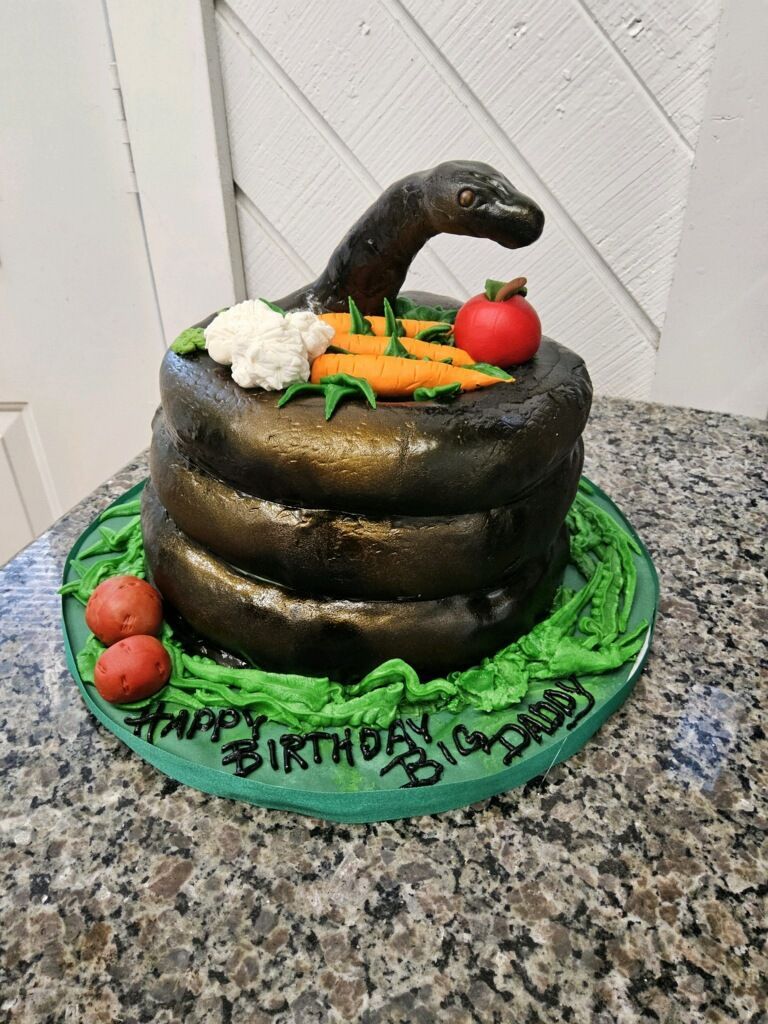 Snake-themed birthday cake with vegetables. Black snake on tiered cake, green frosting base, and text.