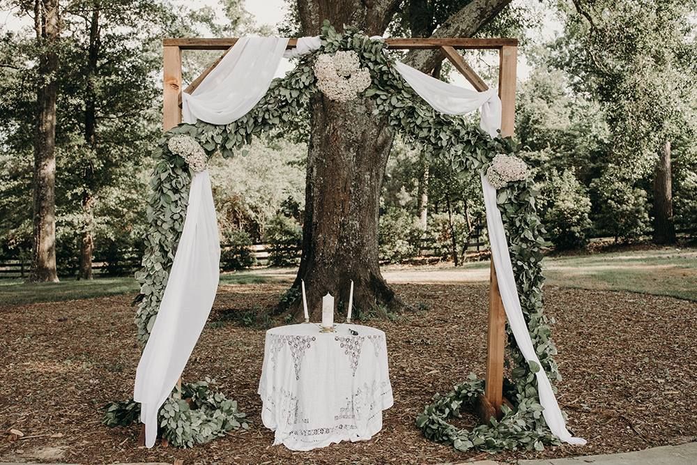 Wedding ceremony setup: Wooden arch draped with white fabric, decorated with greenery and flowers. Table with candles.