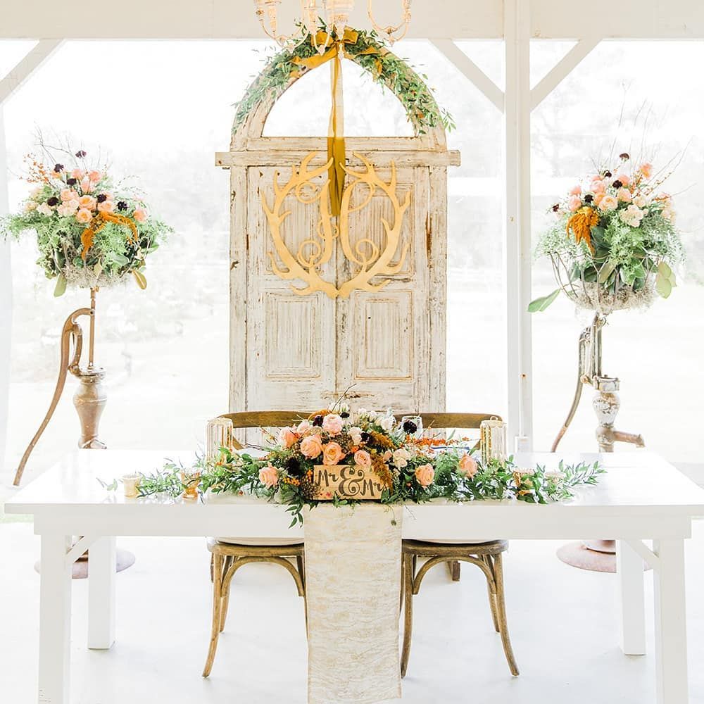 Wedding reception setup: white table with floral arrangements, old door backdrop, and gold chandelier.