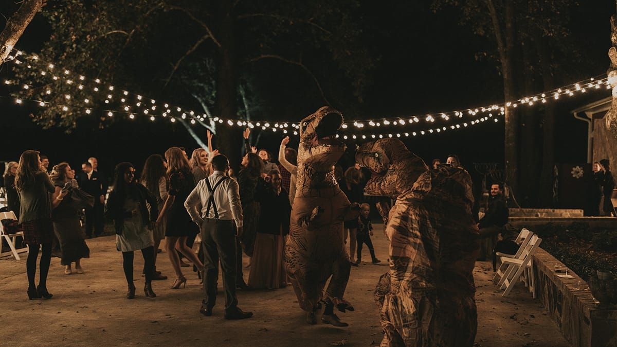 People dancing at an outdoor party at night, including several in dinosaur costumes, under string lights.