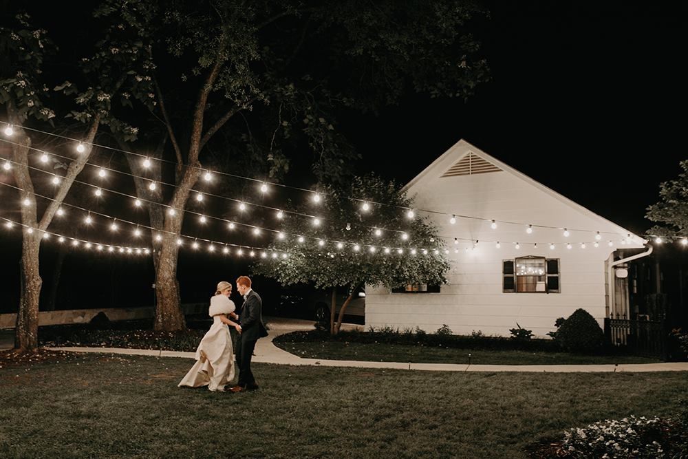 Couple dancing under string lights in front of a white building at night.