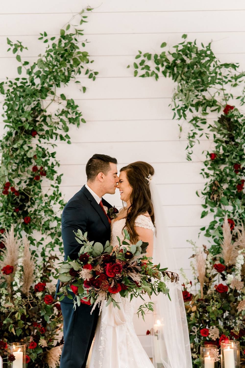 Bride and groom embrace, red floral bouquet. Green archway with red roses. White wall backdrop.