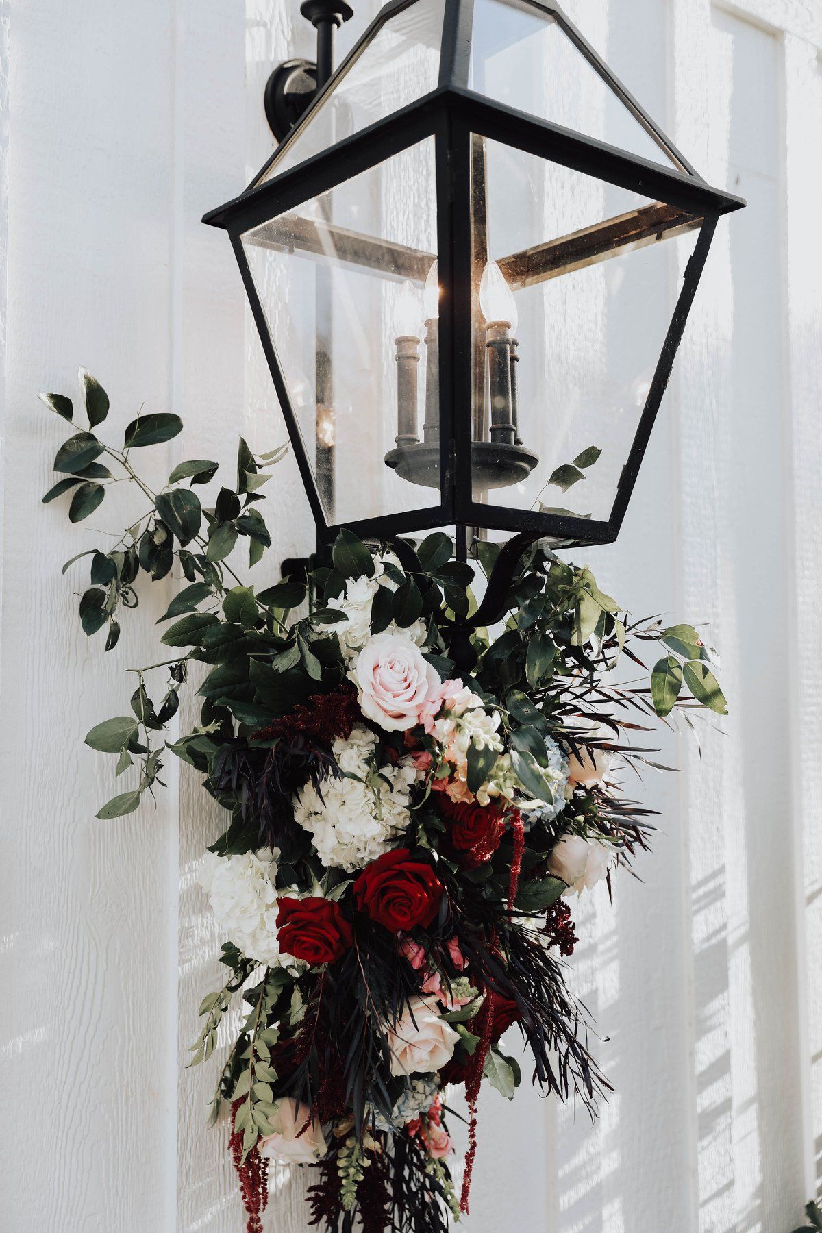 Lantern decorated with cascading floral arrangement; white, red, and green flowers, on a white wall.