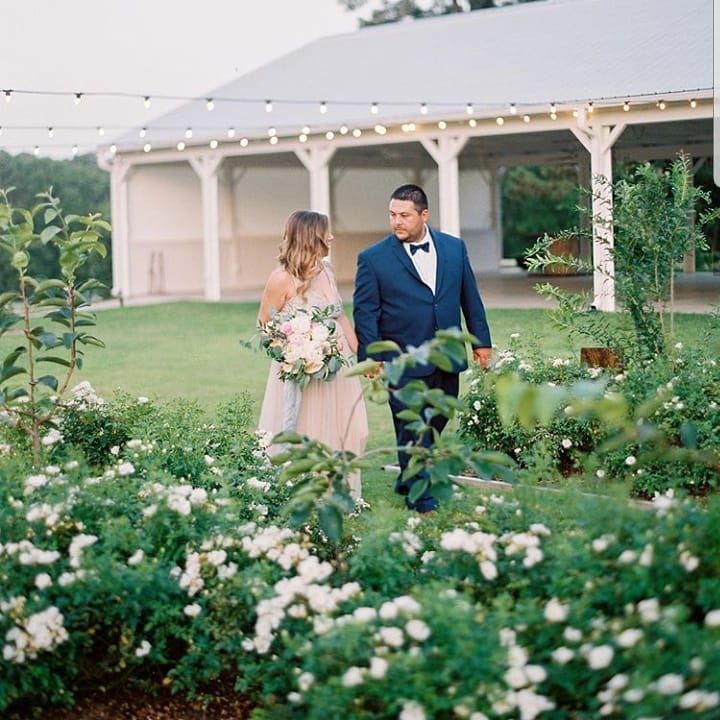 Bride and groom walk hand in hand through a flower garden toward a white gazebo.
