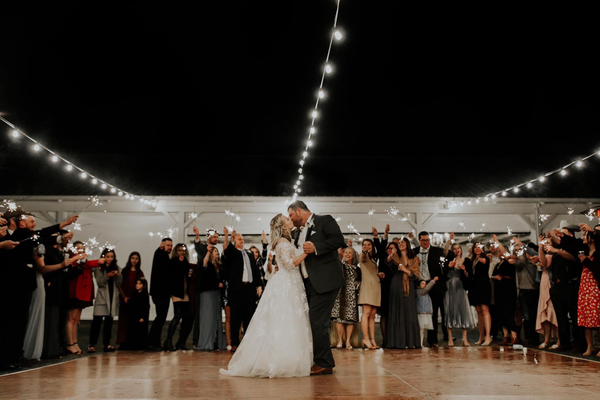 Newlyweds share a kiss on a dance floor, guests cheer and wave sparklers beneath string lights.