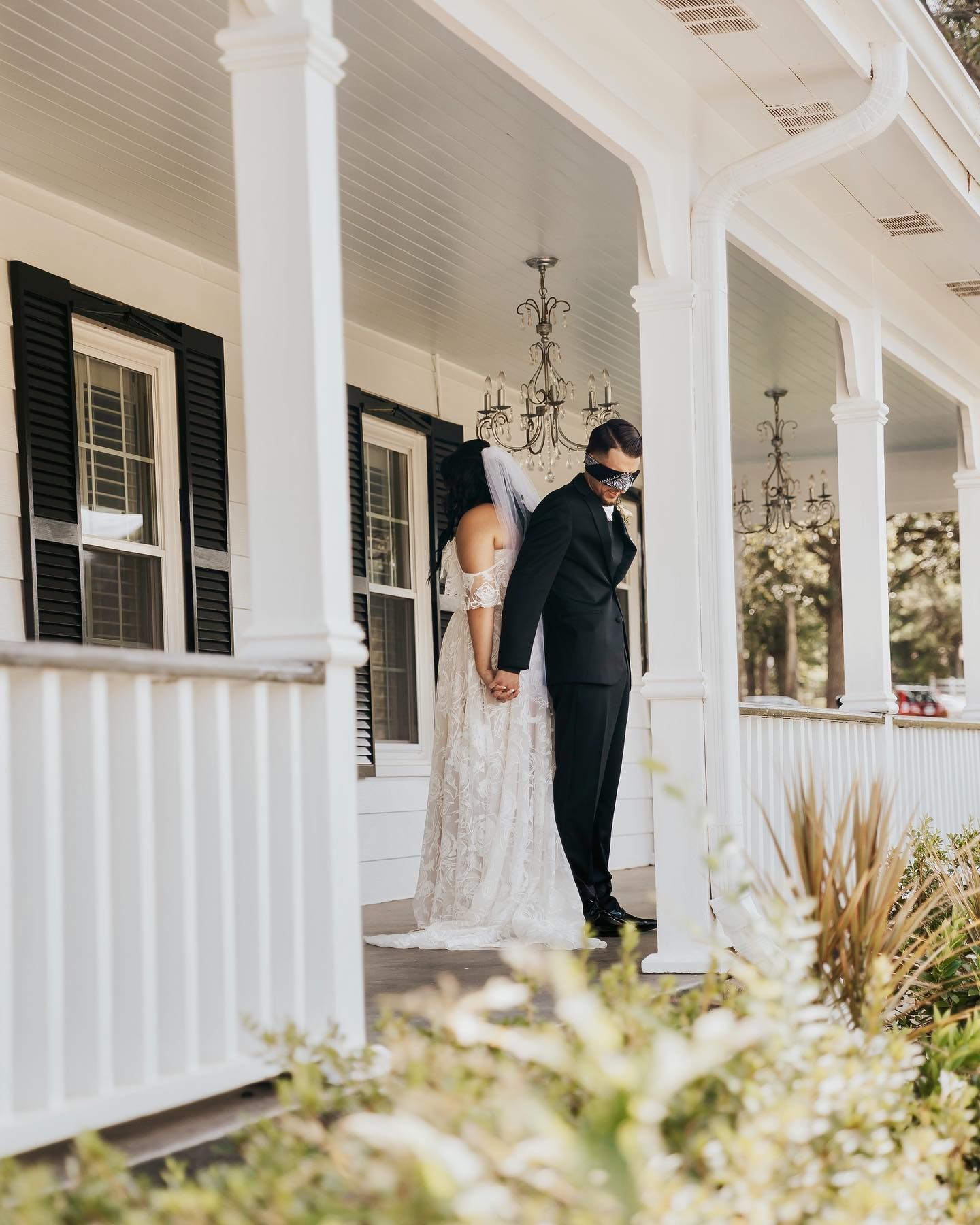 Bride and groom holding hands, blindfolded groom awaiting bride on white porch.