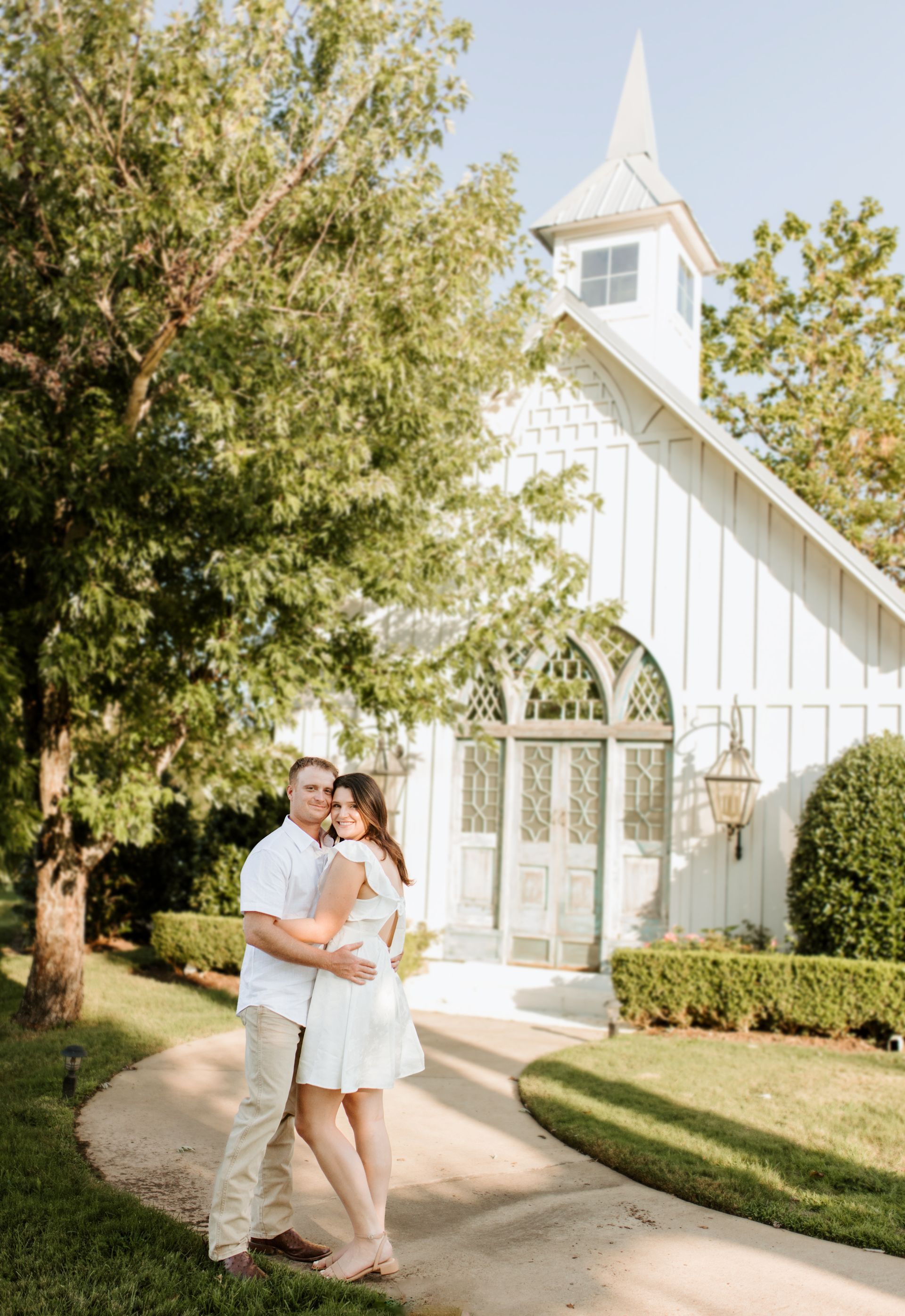 Couple hugging in front of a white chapel; they are on a paved path, sunny day.