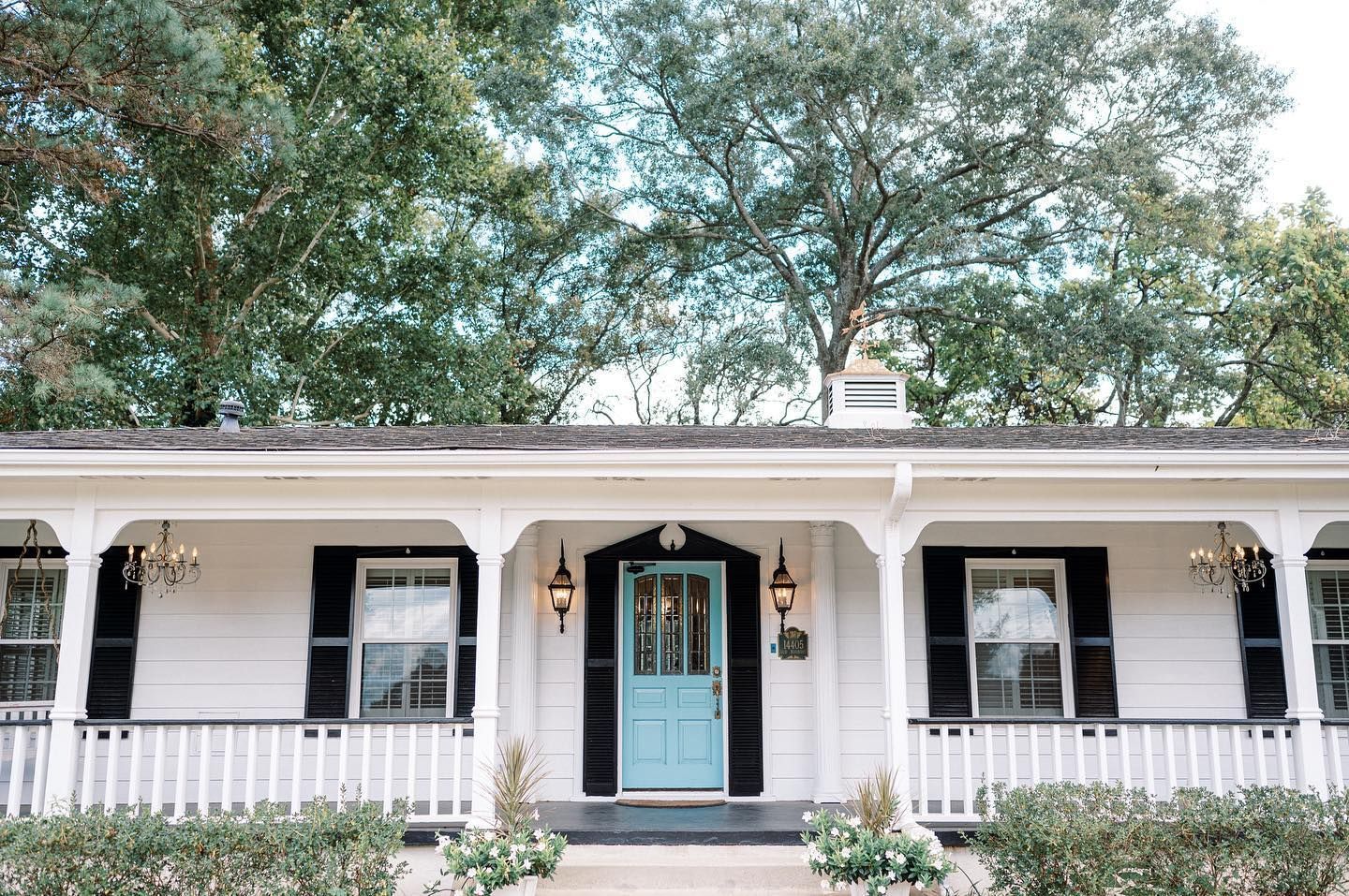 White house with a teal front door, black shutters, and hanging lanterns under a porch; set amongst lush greenery.