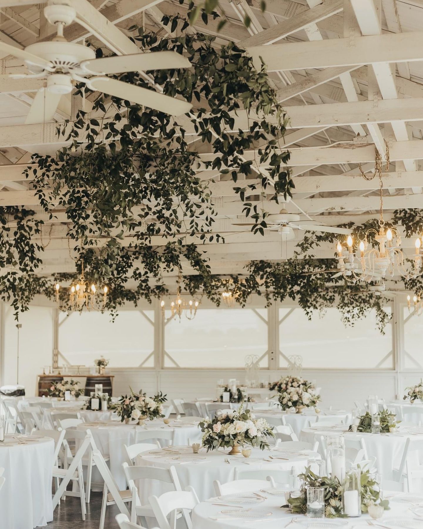 White wedding reception venue with floral arrangements, greenery, and chandeliers.