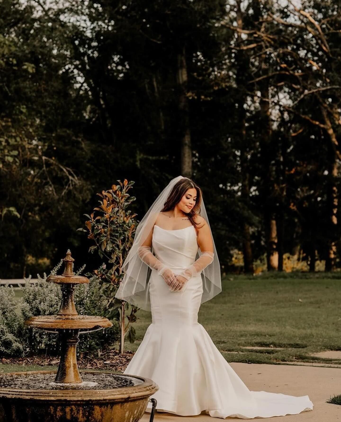 Bride in a white strapless mermaid gown poses near a fountain with veil and gloves outdoors.