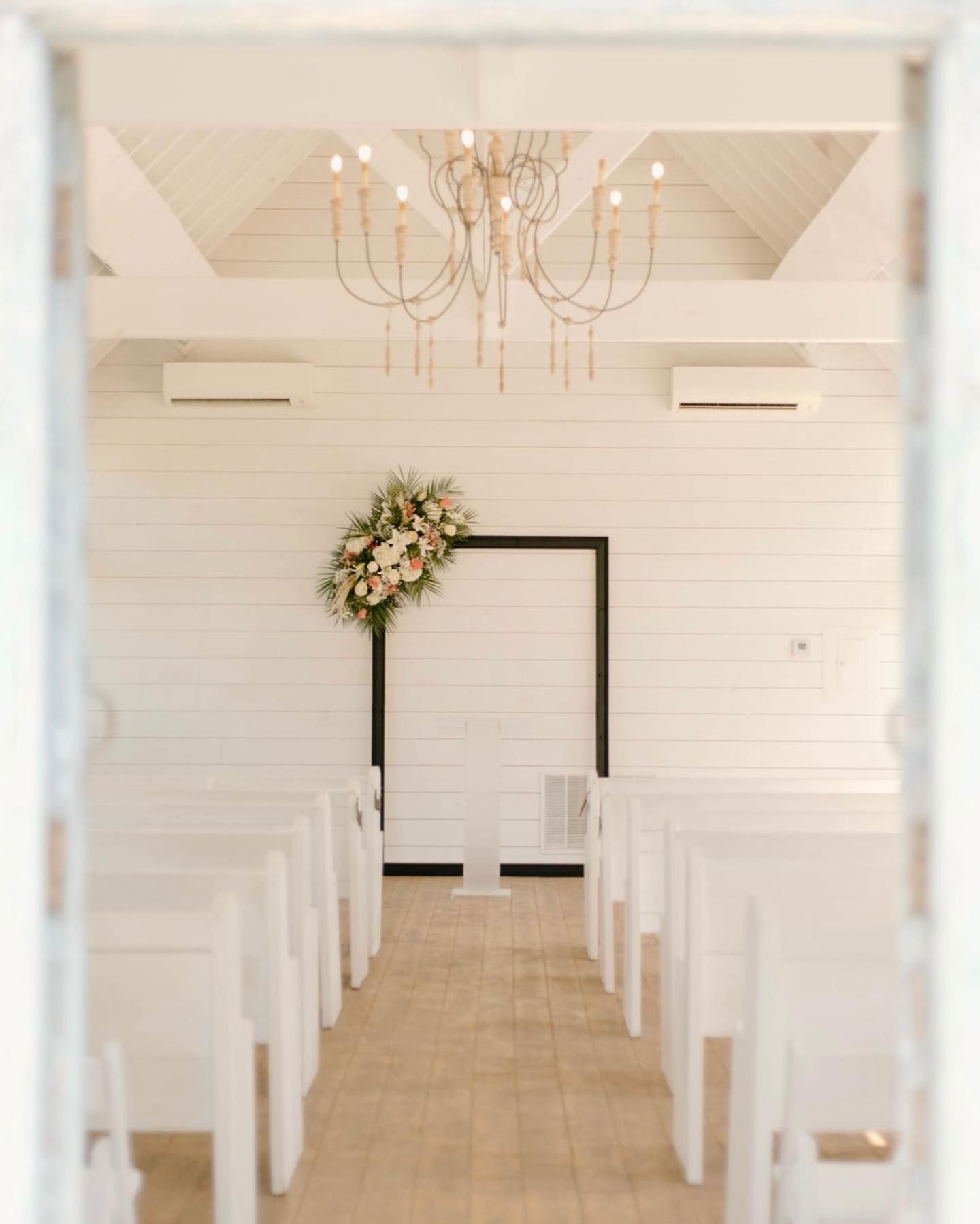 Wedding chapel interior with white walls, pews, floral arrangement, and chandelier.